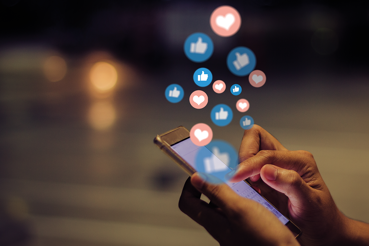 Social Media Marketing: Hände halten ein Handy, aus dem Icons wie ein hochgereckter Daumen und Herzen aufsteigen